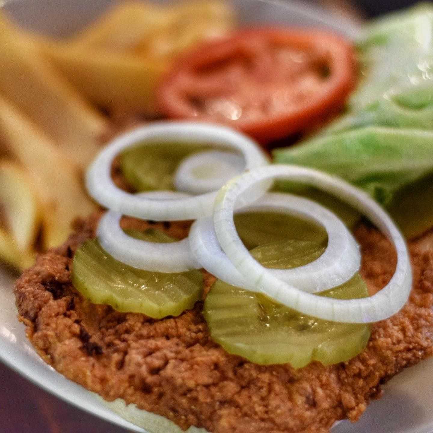 Close-up of a crispy chicken sandwich with pickles, onions, lettuce, tomato, and fries on the side.